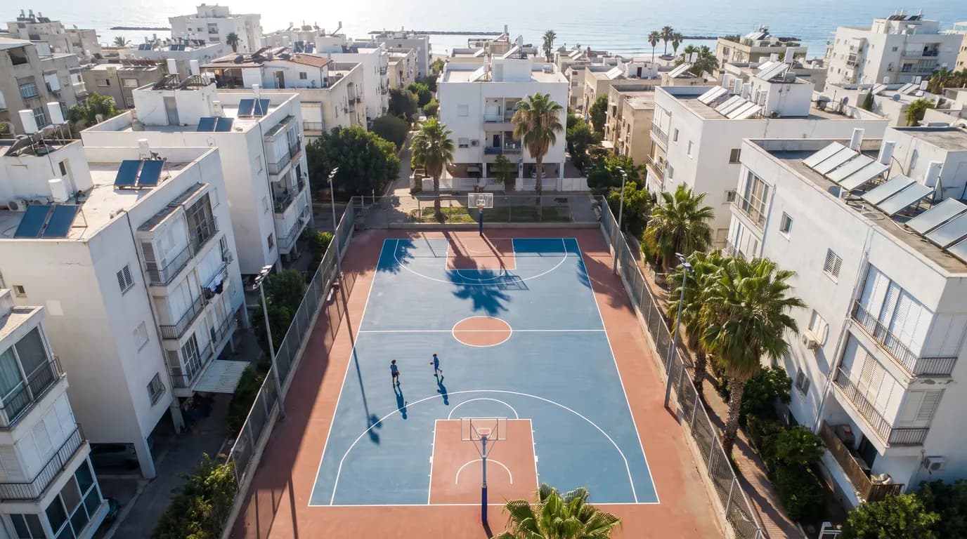 מבט רחפן מלמעלה על מגרש כדורסל שכונתי בישראל עם קווים לבנים טריים, צללים ארוכים של ילדים ועצי דקל באור אחר צהריים