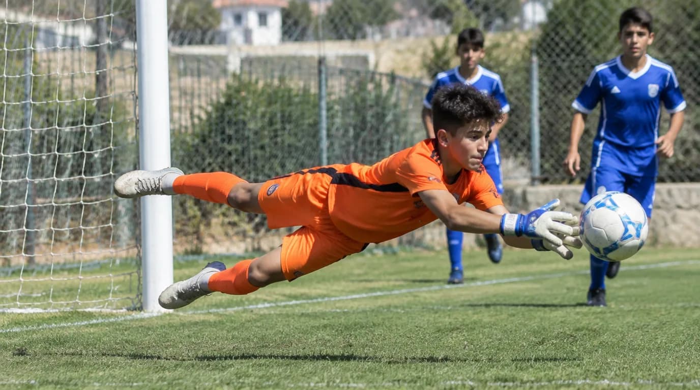 שוער כדורגל ישראלי צעיר לובש חולצה בצבע שונה מהקבוצה, קופץ להדוף כדור ברחבת עונשין במגרש קהילתי
