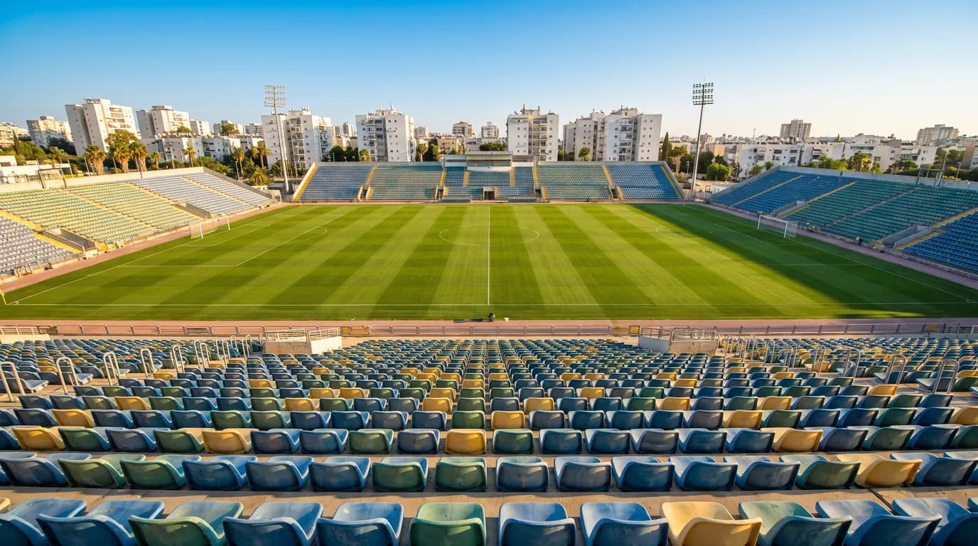 מבט מהיציע על מגרש כדורגל באצטדיון בישראל, עם פסי דשא וקו רקיע עירוני ודקלים ביום שמש